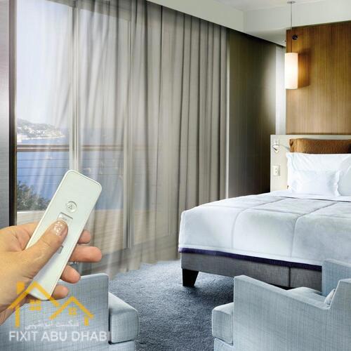 A hand holding a remote control device in a hotel room with large curtains and windows.